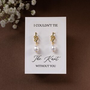 May include: Gold-colored knot earrings with a white pearl drop, displayed on a white card with the text "I Couldn't Tie The Knot Without You". The card is set against a brown background, with white flowers in the upper left corner.