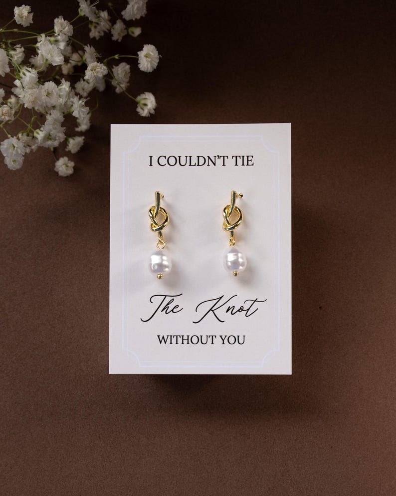 May include: Gold-colored knot earrings with a white pearl drop, displayed on a white card with the text "I Couldn't Tie The Knot Without You". The card is set against a brown background, with white flowers in the upper left corner.