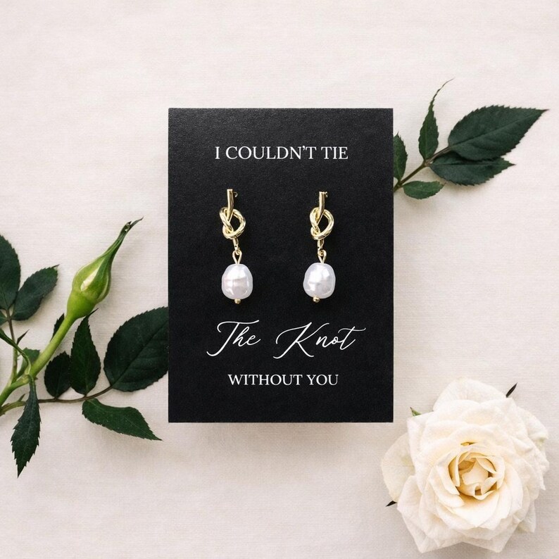 May include: Gold-colored knot earrings with white pearl drops displayed on a black card. The card reads "I COULDN'T TIE The Knot WITHOUT YOU" in elegant script. The image is set against a soft, neutral background with floral accents.