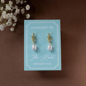 May include: A pair of gold-colored knot earrings with white pearl drops, presented on a light blue card. The card reads "I COULDN'T TIE The Knot WITHOUT YOU" in elegant script. The background is a warm brown.