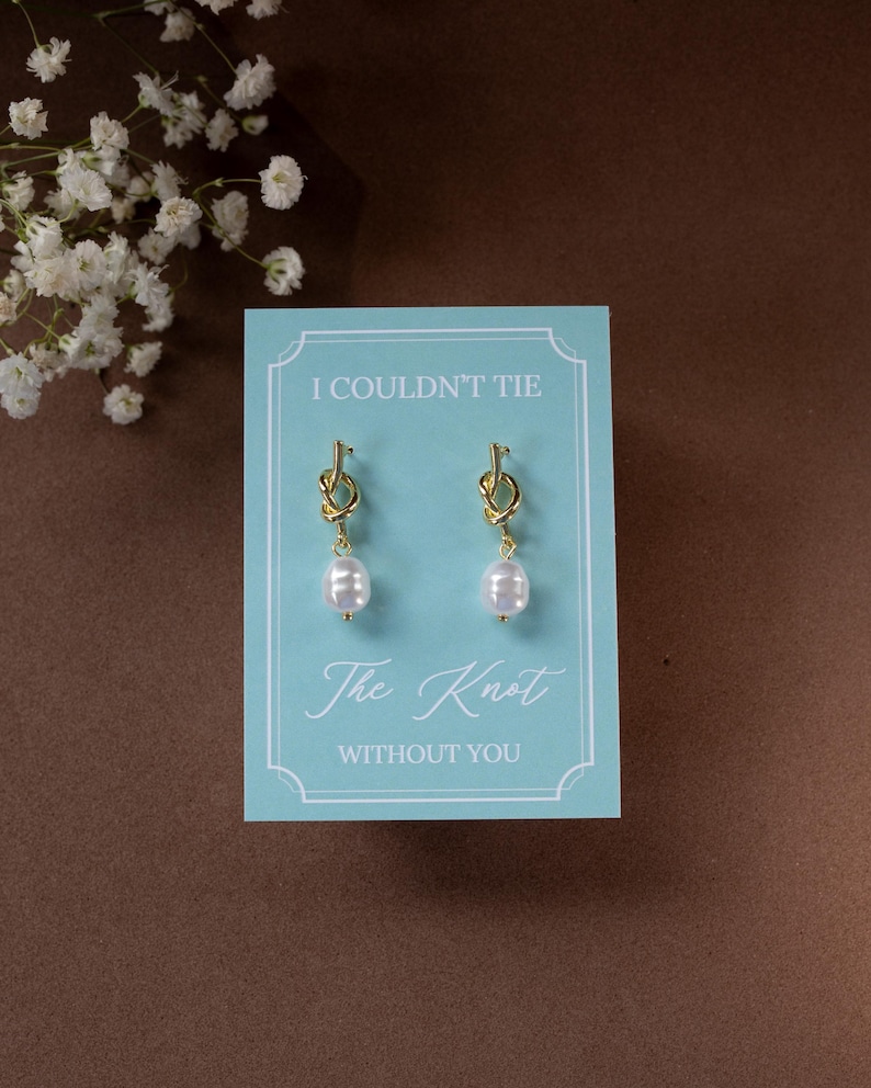 May include: A pair of gold-colored knot earrings with white pearl drops, presented on a light blue card. The card reads "I COULDN'T TIE The Knot WITHOUT YOU" in elegant script. The background is a warm brown.