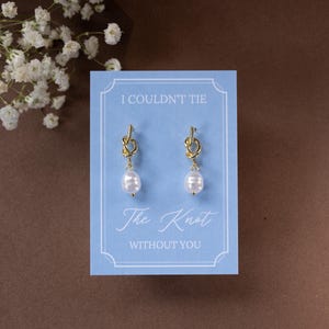 May include: Gold-colored knot earrings with white pearl drops are displayed on a light blue card. The card reads "I COULDN'T TIE The Knot WITHOUT YOU" in elegant script. The background is a brown surface.