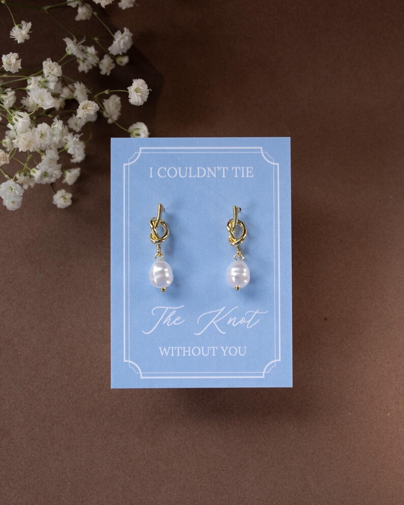 May include: Gold-colored knot earrings with white pearl drops are displayed on a light blue card. The card reads "I COULDN'T TIE The Knot WITHOUT YOU" in elegant script. The background is a brown surface.