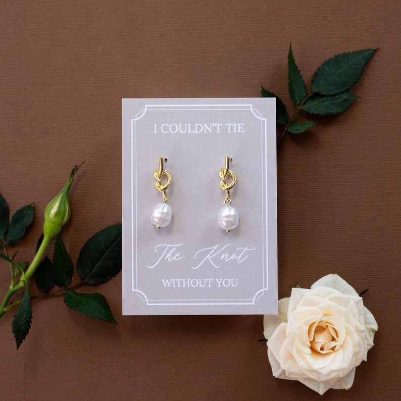 May include: Gold-tone knot earrings with white pearl drops displayed on a gray card. The card reads "I COULDN'T TIE The Knot WITHOUT YOU" in elegant script. A white rose and greenery accent the brown background.