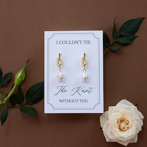 May include: Gold-tone knot earrings with a dangling white pearl, presented on a white card with the text "I COULDN'T TIE The Knot WITHOUT YOU". The card is set against a brown background, with a rose and greenery.