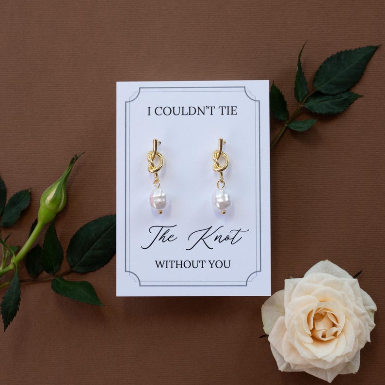 May include: Gold-tone knot earrings with a dangling white pearl, presented on a white card with the text "I COULDN'T TIE The Knot WITHOUT YOU". The card is set against a brown background, with a rose and greenery.