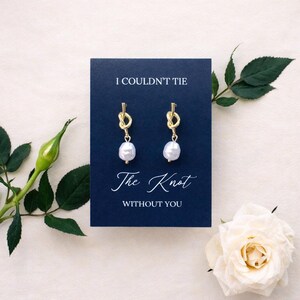 May include: Gold-colored knot earrings with white pearl drops are displayed on a navy blue card that reads "I COULDN'T TIE The Knot WITHOUT YOU". The image includes a white rose and green foliage.