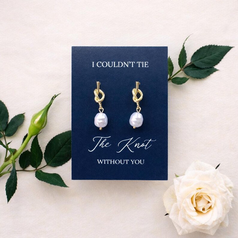 May include: Gold-colored knot earrings with white pearl drops are displayed on a navy blue card that reads "I COULDN'T TIE The Knot WITHOUT YOU". The image includes a white rose and green foliage.