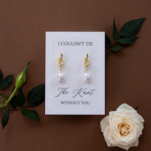 May include: Gold-colored knot earrings with a dangling pearl, presented on a white card with the text "I Couldn't Tie The Knot Without You." The card is set against a brown background, with a white rose and greenery.
