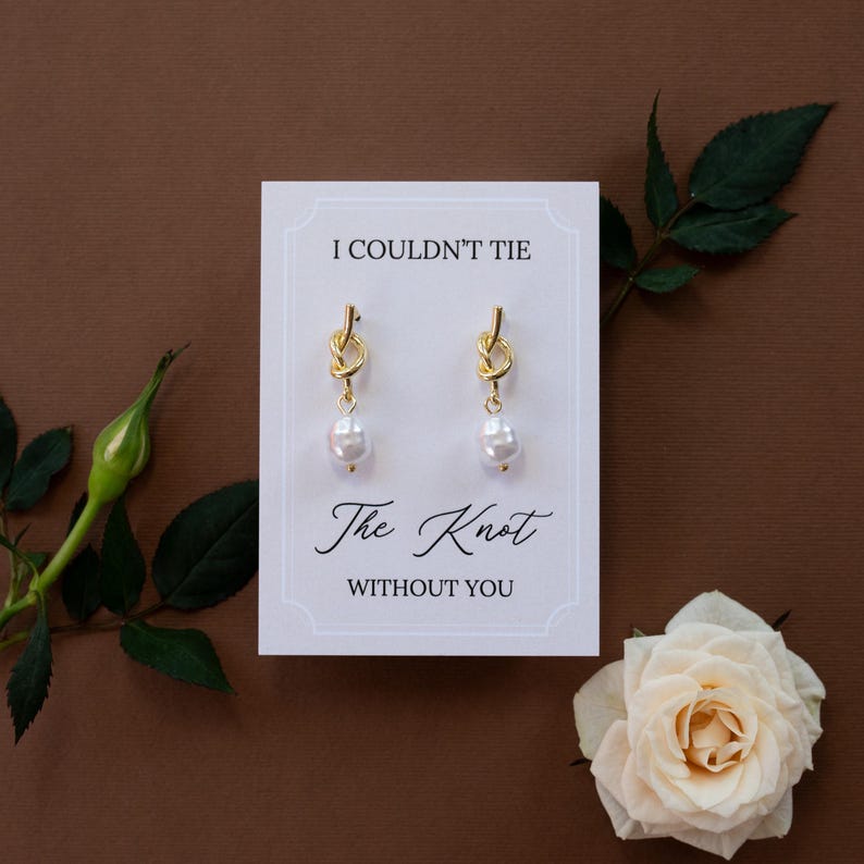 May include: Gold-colored knot earrings with a dangling pearl, presented on a white card with the text "I Couldn't Tie The Knot Without You." The card is set against a brown background, with a white rose and greenery.