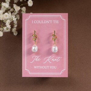 May include: A pair of gold-colored knot earrings with white pearl drops, displayed on a pink card. The card reads "I Couldn't Tie The Knot WITHOUT YOU" in elegant script. The background is a soft brown.