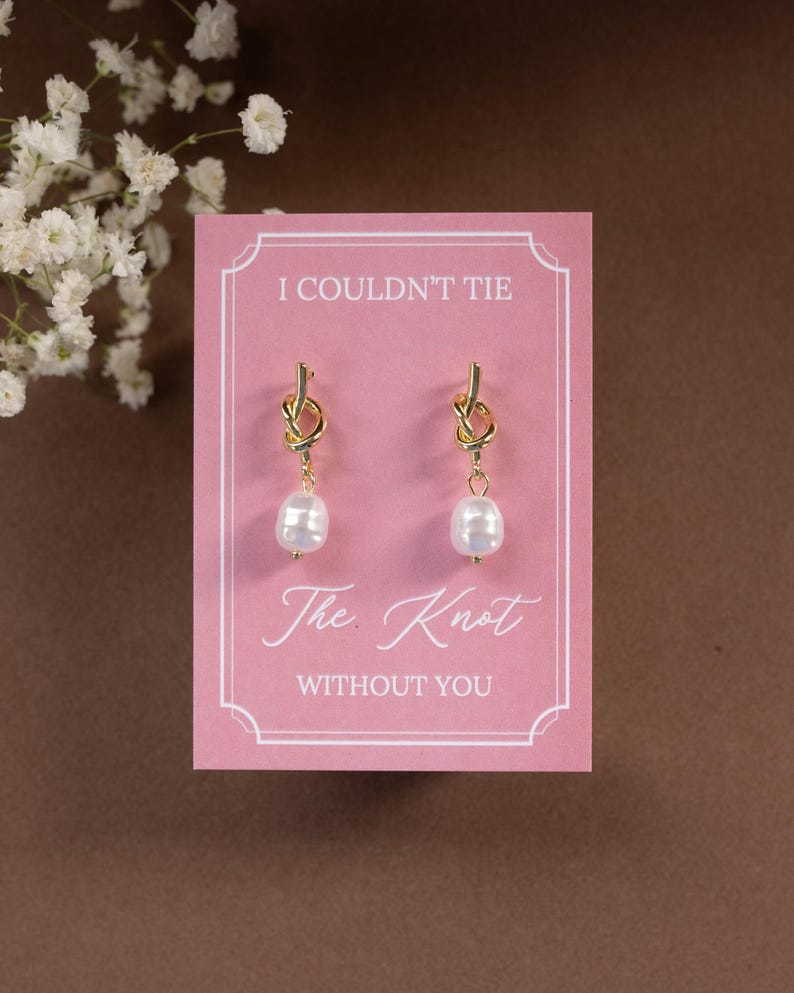 May include: A pair of gold-colored knot earrings with white pearl drops, displayed on a pink card. The card reads "I Couldn't Tie The Knot WITHOUT YOU" in elegant script. The background is a soft brown.