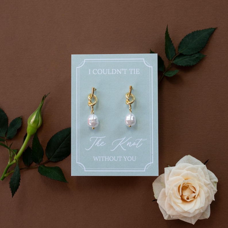 May include: Gold-tone knot earrings with teardrop pearls, displayed on a light blue card with the text "I Couldn't Tie The Knot Without You." The earrings are presented with a white rose and greenery on a brown background.
