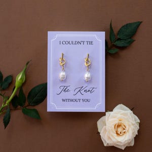 May include: Gold-tone knot earrings with a dangling white pearl, presented on a lavender card. The card reads "I Couldn't Tie The Knot WITHOUT YOU". A white rose and greenery are in the background.