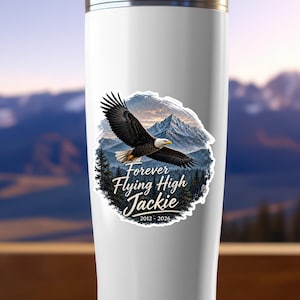 May include: A white cylindrical tumbler featuring a graphic of a bald eagle in flight over a mountain range and pine forest. The text reads Forever Flying High Jackie 2012 - 2026 in a white script font centered below the eagle illustration.