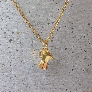 Peut inclure: Un collier doré avec un petit pendentif en forme de cafetière. Le charme est suspendu à une fine chaîne dorée. L'arrière-plan est une vue floue d'une surface en béton gris et d'une plante verte.