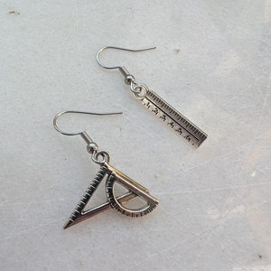 Op de afbeelding: Een paar zilveren oorbellen met geometrische vormen. Eén oorbel heeft een liniaal met markeringen van 1 tot 8. De andere oorbel heeft een gradenboog en een driehoek.