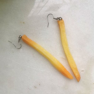 Op de afbeelding: Een paar oorbellen in de vorm van frietjes. De oorbellen zijn gemaakt van geel en oranje plastic en hebben zilveren haakjes.