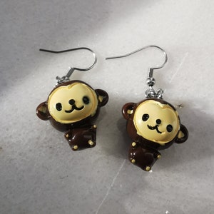 Puede incluir: Un par de pendientes de mono marrones y amarillos con ganchos plateados. Los monos tienen una cara de dibujos animados linda con ojos negros y una sonrisa.