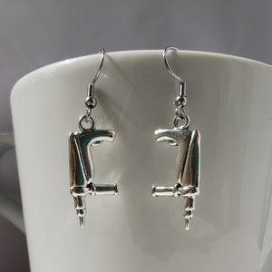 May include: A pair of silver earrings shaped like a drill. The earrings are hanging from a white mug.