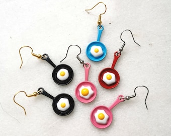 May include: Five pairs of dangle earrings featuring miniature frying pans with a fried egg design. The pans are in various colors, including blue, red, black, and pink.