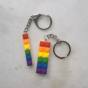 Può includere: Due portachiavi in ​​mattoncini Lego arcobaleno attaccati a portachiavi argentati. Il portachiavi a sinistra è attaccato a un moschettone argentato. Il portachiavi a destra è attaccato a un anello diviso argentato.