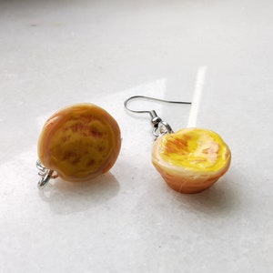 Puede incluir: Pendientes hechos a mano con un diseño de melocotón en miniatura. Los pendientes tienen un aspecto realista con tonos de amarillo, naranja y rojo. Los pendientes están sujetos a ganchos para las orejas de color plateado.