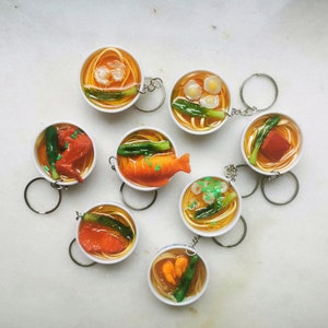 May include: A set of eight colorful keychains shaped like bowls of ramen noodles. Each keychain features a different topping, including eggs, fish, and vegetables.