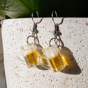 May include: A pair of silver dangle earrings with a clear glass beer mug charm. The mug is filled with a yellow liquid and has a white foam head.