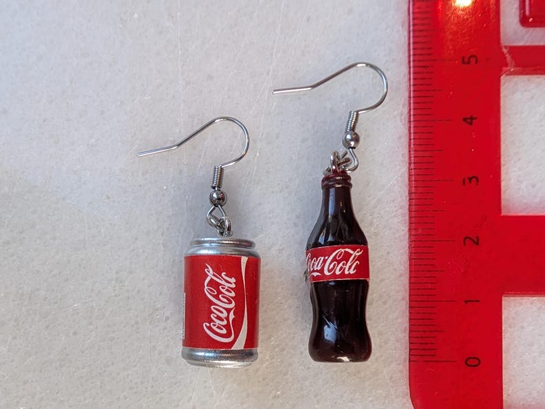 Puede incluir: Un par de pendientes de plata con un encanto de lata y botella de Coca-Cola. La lata es plateada con la marca Coca-Cola roja y blanca. La botella es marr&oacute;n con una etiqueta de Coca-Cola roja y blanca.