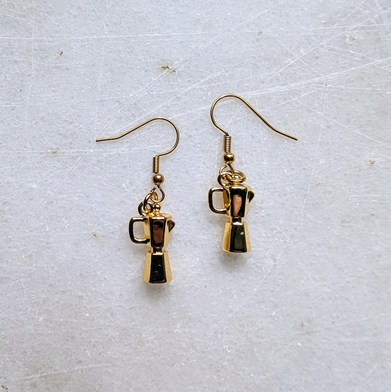 May include: A pair of gold-colored earrings featuring miniature moka pot charms. The earrings have a simple hook design and are suspended from a small gold bead. The moka pot charms have a detailed, three-dimensional design.