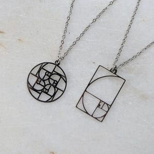 Puede incluir: Dos collares con colgantes geométricos. Un colgante es un círculo negro calado con un diseño en espiral. El otro es un rectángulo negro calado con un diseño en espiral. Ambos colgantes cuelgan de cadenas plateadas.