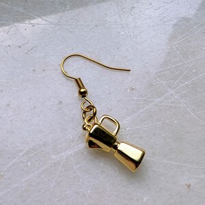May include: A single gold-colored earring featuring a miniature coffee maker charm. The charm has a rectangular base, a cylindrical body, and a handle. The earring hook is also gold and has a simple, curved design. The background is a light, textured surface.