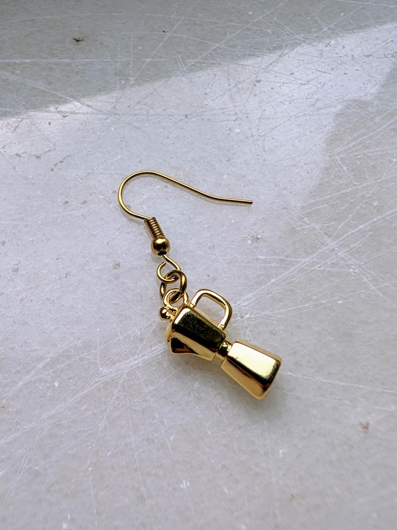 May include: A single gold-colored earring featuring a miniature coffee maker charm. The charm has a rectangular base, a cylindrical body, and a handle. The earring hook is also gold and has a simple, curved design. The background is a light, textured surface.