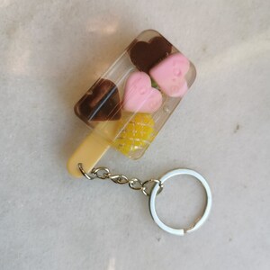 以下が含まれることがあります： 黄色のパイナップル、ピンクのハート2つ、茶色のチョコレートのハート2つが透明なプラスチック製のアイスキャンディーの形をしたキーホルダー。キーホルダーには銀色の金属製のリングが付いています。
