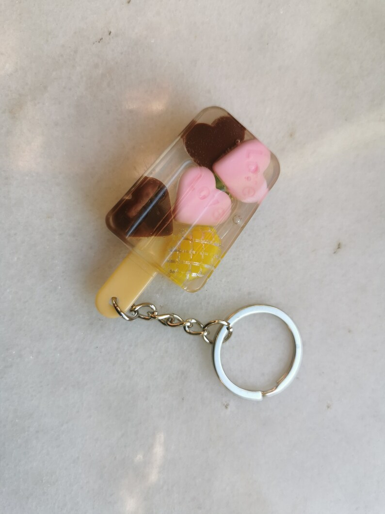 以下が含まれることがあります： 黄色のパイナップル、ピンクのハート2つ、茶色のチョコレートのハート2つが透明なプラスチック製のアイスキャンディーの形をしたキーホルダー。キーホルダーには銀色の金属製のリングが付いています。