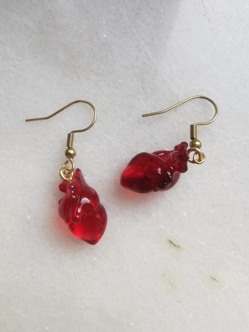May include: A pair of gold heart-shaped earrings with red glass hearts. The earrings are dangling from gold hooks.
