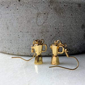 May include: A pair of gold-colored earrings shaped like miniature coffee makers. The earrings have a detailed design, including a spout, handle, and lid. The earrings are displayed against a neutral background.
