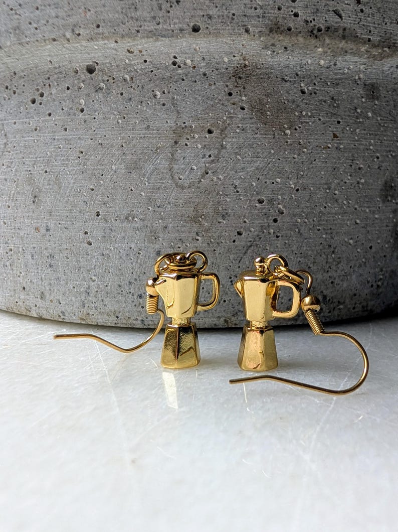 May include: A pair of gold-colored earrings shaped like miniature coffee makers. The earrings have a detailed design, including a spout, handle, and lid. The earrings are displayed against a neutral background.