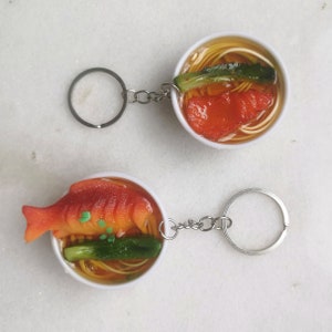 May include: Two keychains shaped like bowls of ramen noodles. The bowls are white with a red fish, green vegetables, and yellow noodles inside. The keychains are attached to silver key rings.
