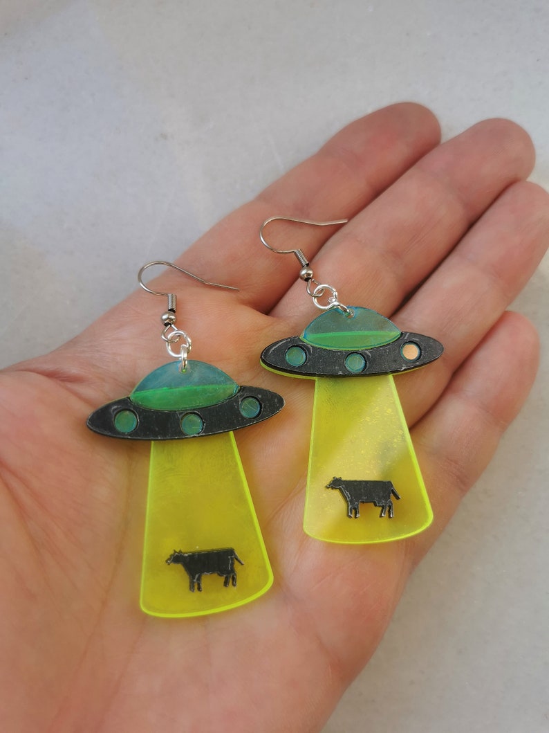 May include: A pair of yellow and green UFO earrings with black cows inside. The earrings are made of acrylic and have silver hooks.