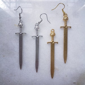 Puede incluir: Dos pares de pendientes con forma de espada. Un par es plateado, el otro dorado. Cada pendiente presenta un diseño de espada con una parte superior decorativa y un gancho para orejas perforadas. Los pendientes se muestran sobre una superficie clara.
