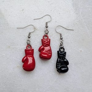 Puede incluir: Tres pares de pendientes con dijes de guantes de boxeo. Un par tiene guantes de boxeo negros, y los otros dos pares tienen guantes de boxeo rojos.