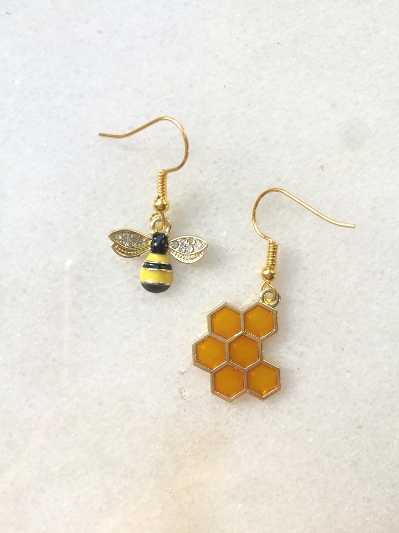 K&ouml;nnte beinhalten: Ein Paar goldene Ohrringe mit einem Bienen- und Wabenmuster. Die Biene ist gelb und schwarz mit klaren Strasssteinen. Die Wabe ist gelb und gold.