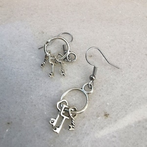 May include: A pair of silver key earrings with three small keys dangling from each earring. The earrings are on a white background.