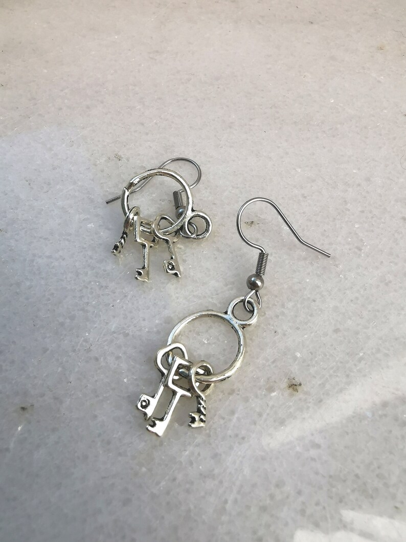 May include: A pair of silver key earrings with three small keys dangling from each earring. The earrings are on a white background.