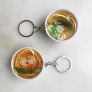 May include: Two white ceramic bowls filled with a simulated noodle soup with shrimp and green vegetables. Each bowl is attached to a silver key chain.