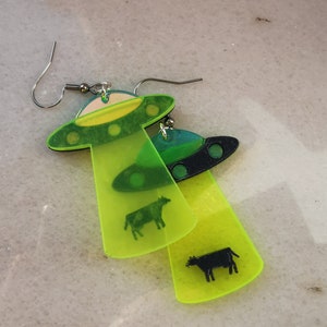 May include: A pair of neon green and black acrylic earrings shaped like UFOs with a cow silhouette inside each one.