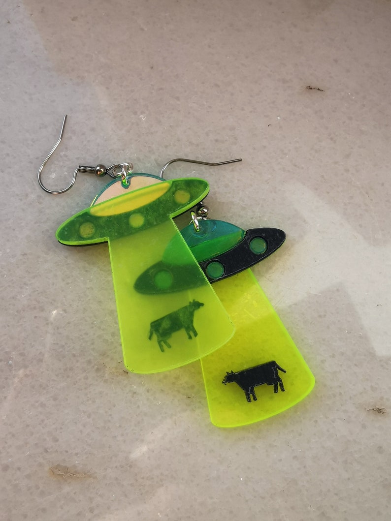 May include: A pair of neon green and black acrylic earrings shaped like UFOs with a cow silhouette inside each one.