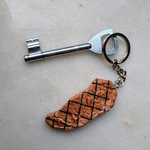 May include: A silver key with a brown and black grilled steak keychain. The steak is a realistic replica of a grilled steak.